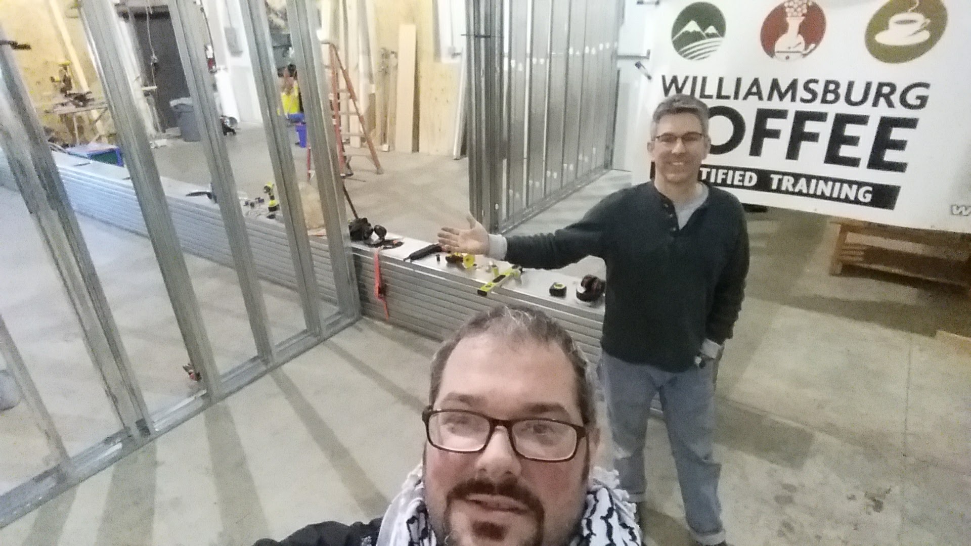 Two men stand taking a selfie in a construction zone with a wall being built in the background and sign for Williamsburg Coffee Certified Training